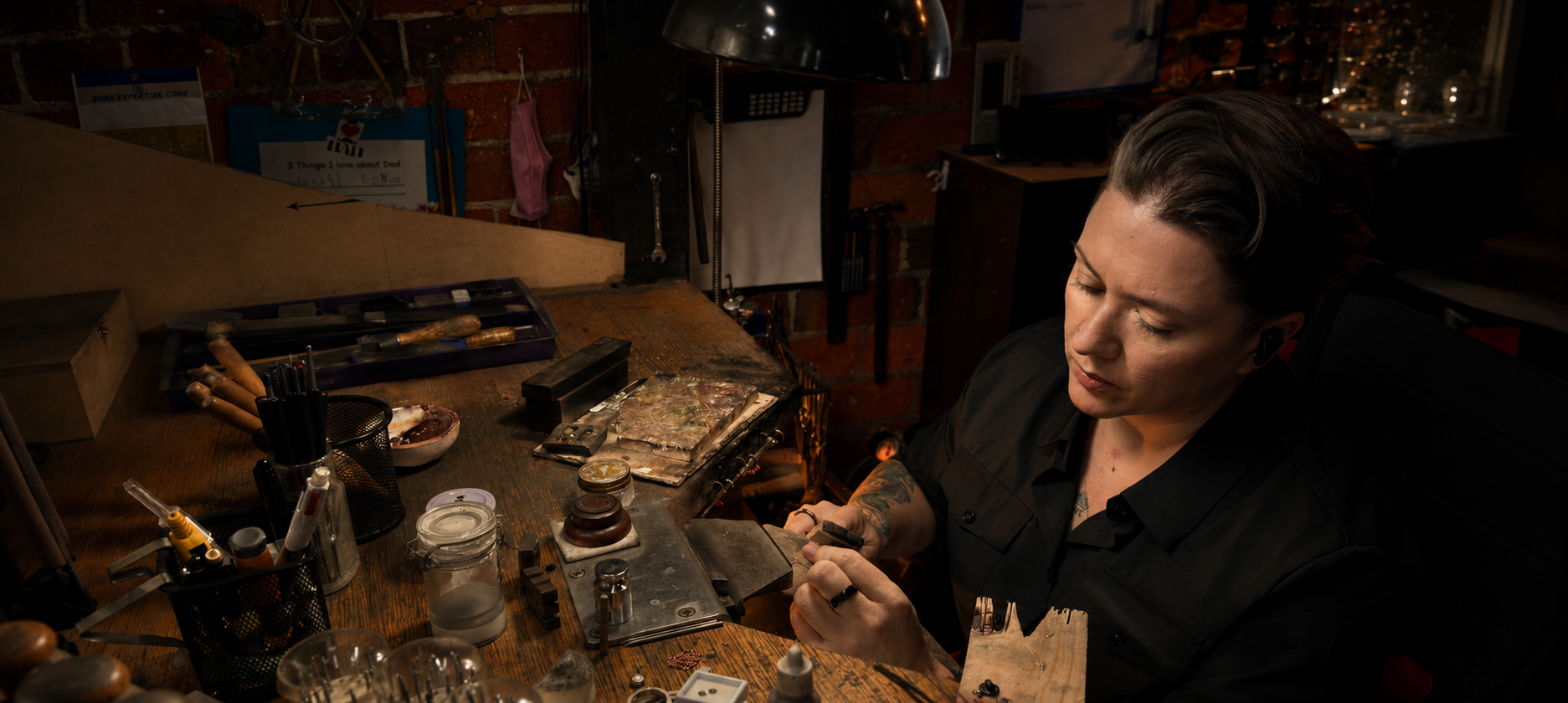 Woman working at a jewellery bench with various items including bottles and tools in a dimly lit room.